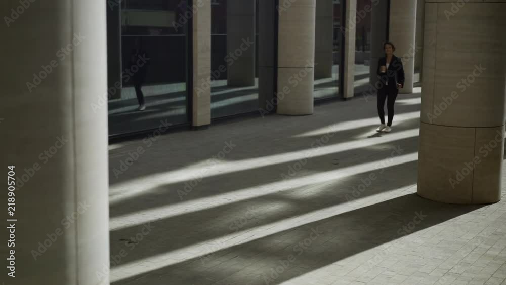 Lockdown shot of elegant middle adult businesswoman wearing casual clothing walking among columns with takeaway coffee cup in her hand and laptop in case under arm Lockdown shot of elegant middle adult businesswoman wearing casual clothing walking among columns with takeaway coffee cup in her hand and laptop in case under arm