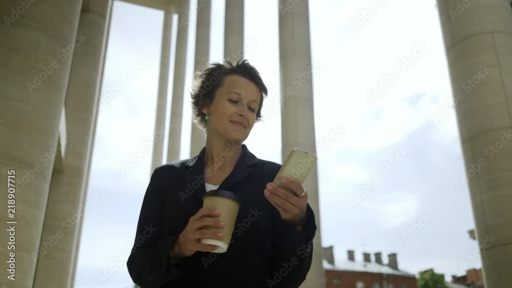 Panning medium shot of confident middle aged businesswoman with short hair texting on cell phone outdoors and smiling, takeaway cup of coffee in her hand Panning medium shot of confident middle aged businesswoman with short hair texting on cell phone outdoors and smiling, takeaway cup of coffee in her hand