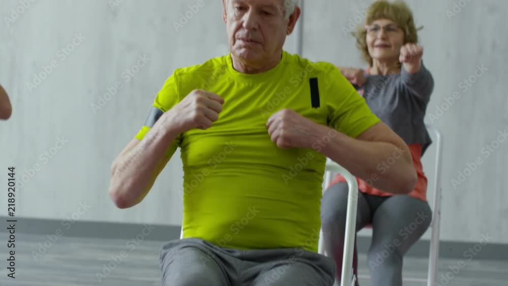 Tilt up of focused senior man and cheerful elderly lady sitting on folding chairs and doing exercises during chair aerobics class