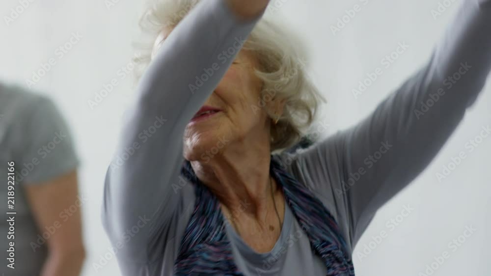 Handheld tracking shot of happy senior woman smiling and swinging raised arms during dynamic stretching group class