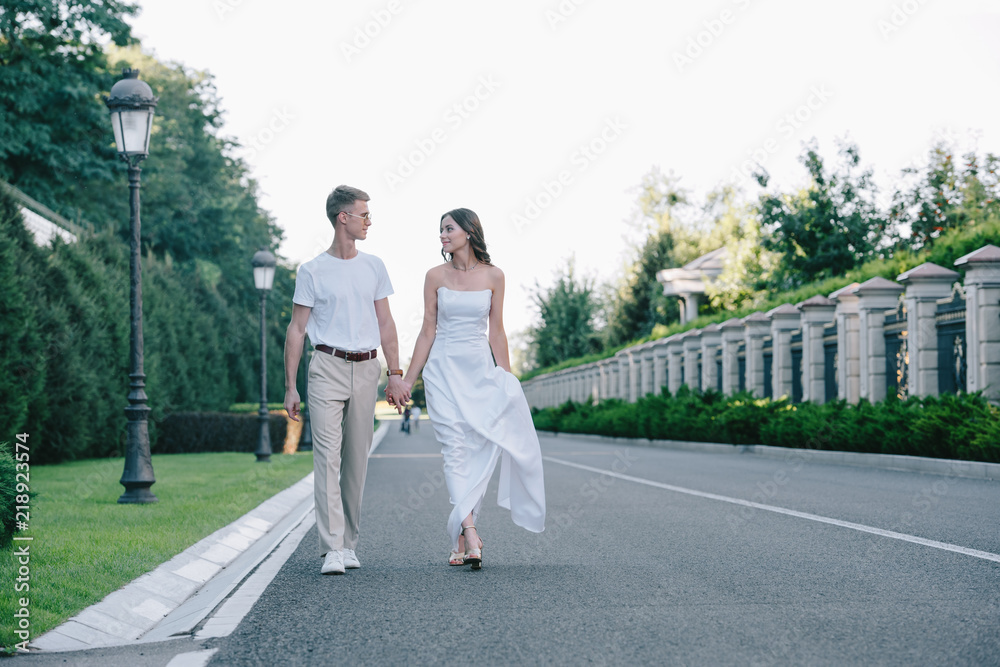 Fototapeta premium bride in wedding dress and groom holding hands and walking on road