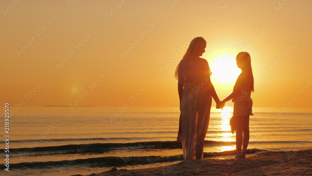 Mom and daughter are looking forward to a beautiful sunset over the sea. Silhouettes of a woman with a child near the water