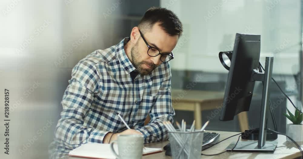 Young Caucasian man in glasses working at the table in his office and writing down something in front of the computer screen.