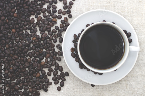 White mug with coffee on a napkin. Spilled coffee beans. Light background, toned photo. Copy space