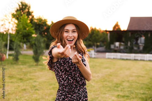 Photo of beautiful young woman 20s wearing dress and straw hat smiling, and inviting to follow her with hand gestures while walking outdoor in countryside