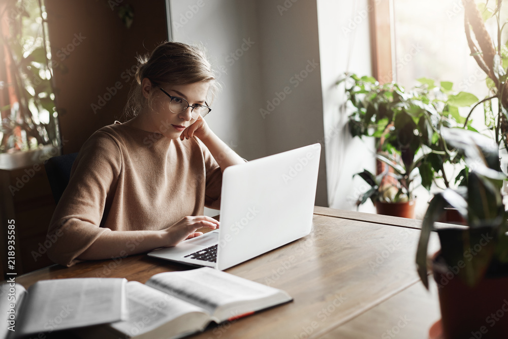 Cute european female coworker in glasses sitting near window in cafe, leaning head on hand and reading articles in network via laptop, searching information for work, making notes in notebook Cute european female coworker in glasses sitting near window in cafe, leaning head on hand and reading articles in network via laptop, searching information for work, making notes in notebook