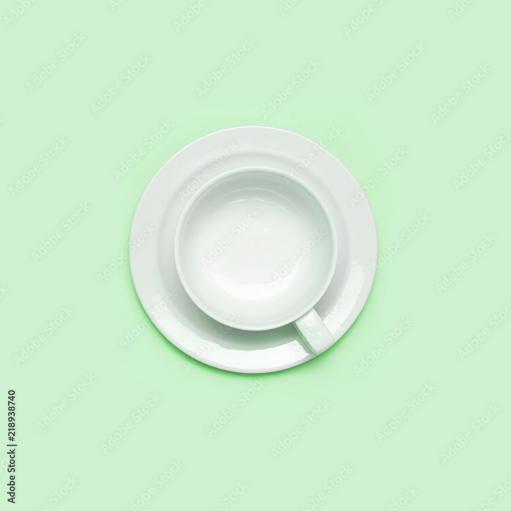 Flat lay top view White empty ceramic cup on a saucer on green background. Concept morning breakfast, drink coffee or tea. Background utensils, kitchen items. Minimalistic background Flat lay top view White empty ceramic cup on a saucer on green background. Concept morning breakfast, drink coffee or tea. Background utensils, kitchen items. Minimalistic background