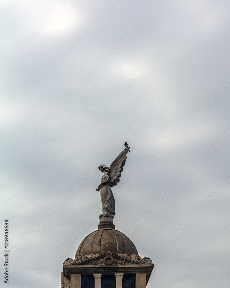 Fototapeta premium Angel sculpture and a magpie against the cloudy sky in Poblenou Cemetery. Cemetery of Poblenou is today home to incredible sculptures, haunting, yet beautiful.