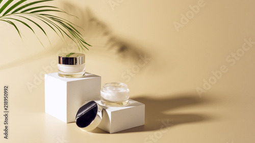 close up view of palm leaf, facial and body creams in glass jars on white cubes on beige background photo