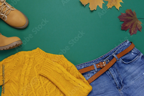 Female orange knitted sweater, blue jeans, boots and autumn leaves on green background top view flat lay. Fashion Lady Clothes Set Trendy Cozy Knit Jumper Autumn accessories Female fashion look