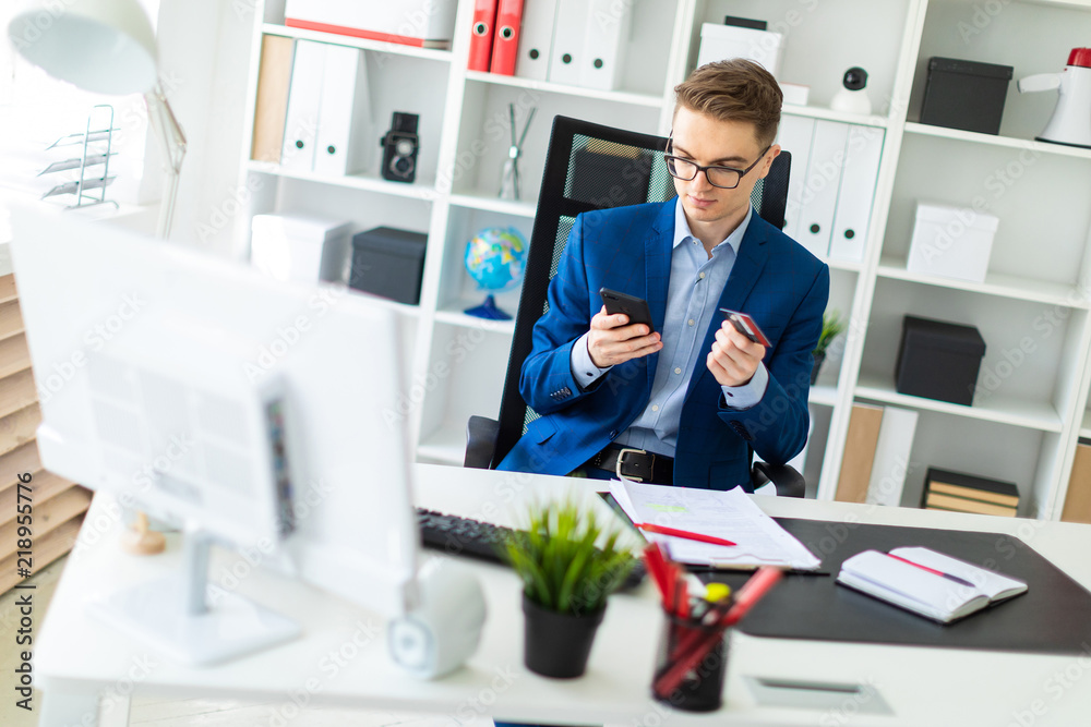 A young man sits at a table in the office and holds a bank card and phone. A young man sits at a table in the office and holds a bank card and phone.