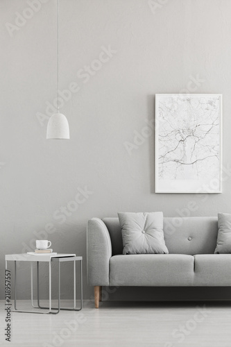 Real photo of map poster hanging on the wall in living room interior with grey sofa, metal end table with books and coffee cup and white lamp