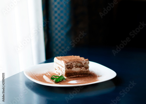 Tiramisu traditional Italian dessert on white plate on dark blue table next to roon window with curtains . Withe day light from the window. Close up food with fork and knife decor