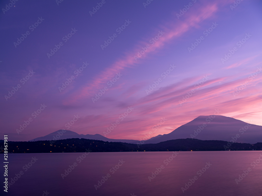夕焼けの湖畔にて、青い空は飛行機雲がピンク色にな り湖面に写る、遠くの山々はシルェツト。