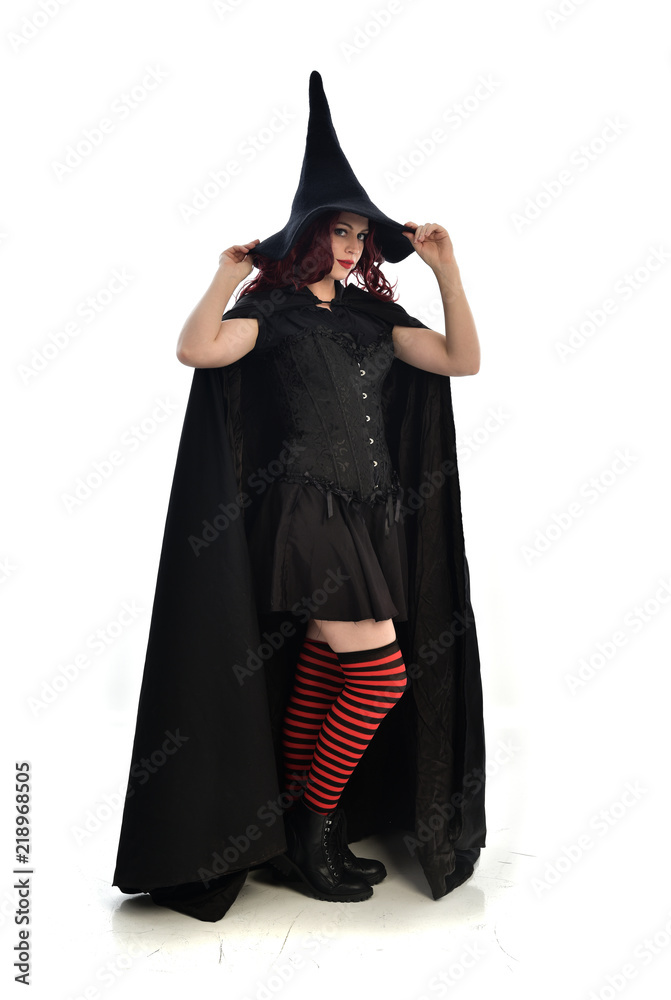 full length portrait of red haired girl wearing long black cloak, pointy hat and witch costume. standing pose, isolated on white studio background. full length portrait of red haired girl wearing long black cloak, pointy hat and witch costume. standing pose, isolated on white studio background.