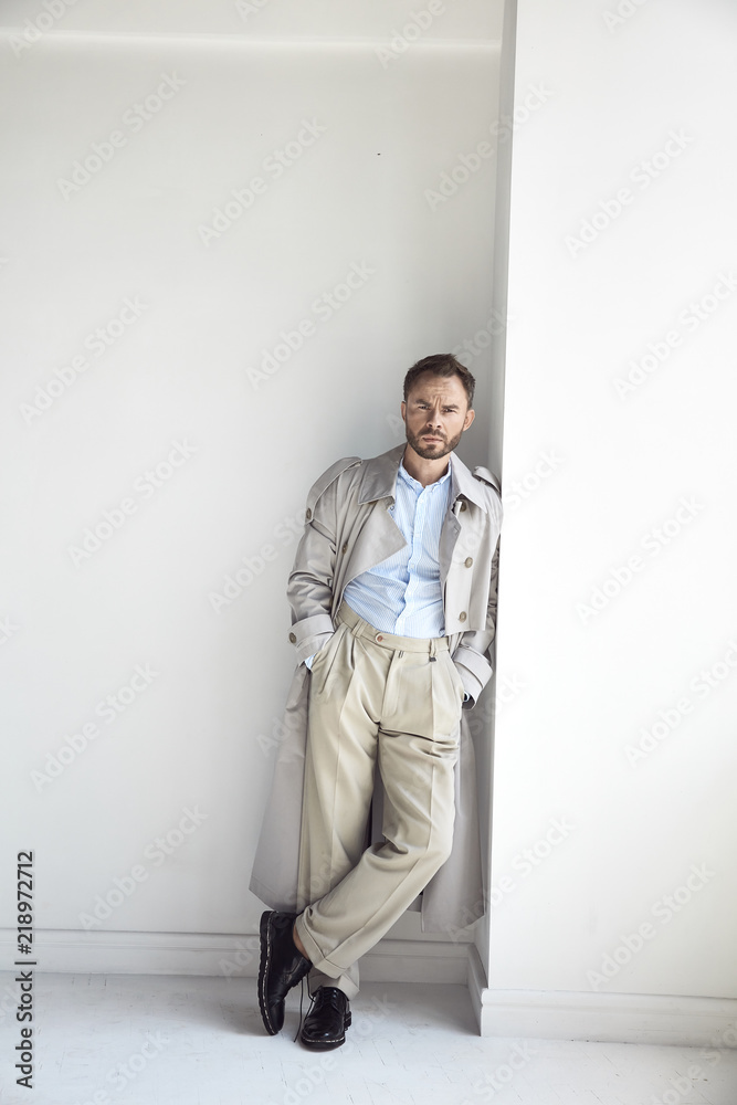 Fashion portrait of handsome man with dark beard and hair, weared in light  trench coat, shirt, beige pants and black shoes. Stock-Foto | Adobe Stock