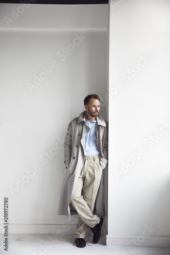 Fashion portrait of handsome man with dark beard and hair, weared in light trench coat, shirt, beige pants and black shoes. Man stands near white wall