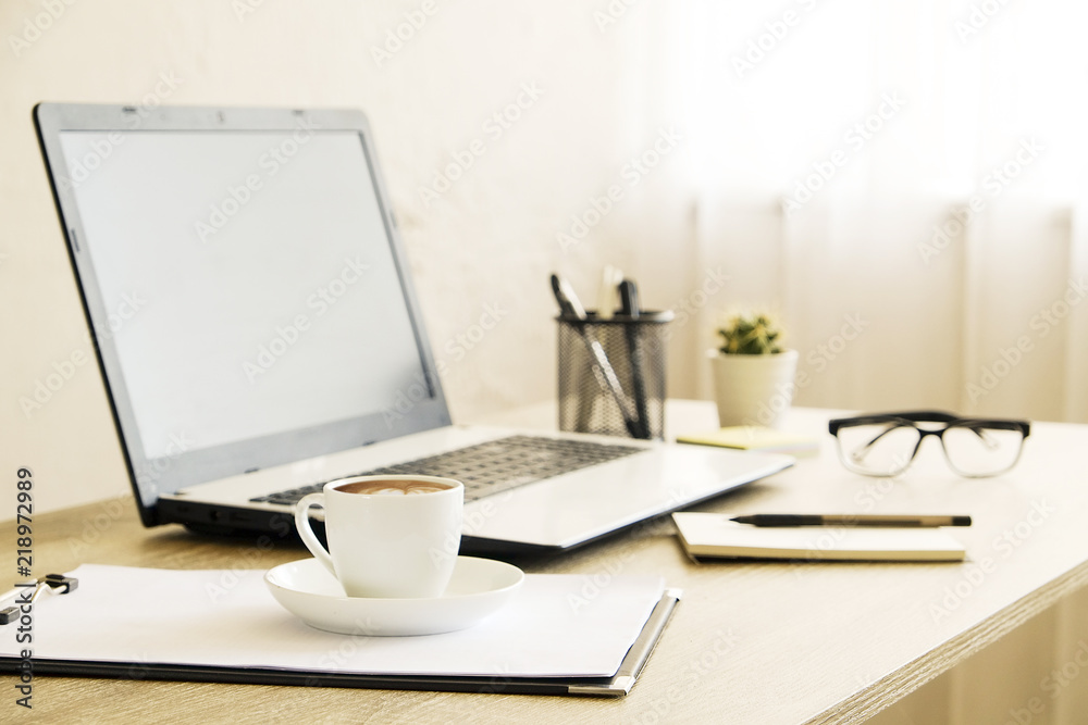 Fototapeta premium Blank screen laptop computer, cup of cappuccino coffee, cactus, supplies and folded eye glasses on wooden desk in spacious office full of sunlight. Creative workspace. Close up, copy space, background