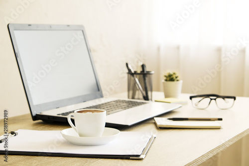 Blank screen laptop computer, cup of cappuccino coffee, cactus, supplies and folded eye glasses on wooden desk in spacious office full of sunlight. Creative workspace. Close up, copy space, background