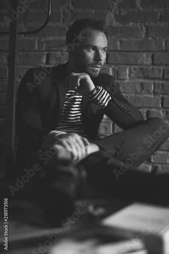 Black and white portrait of handsome stylish man with dark hair and beard in jacket. Businessman at his work place. Brick wall on background photo