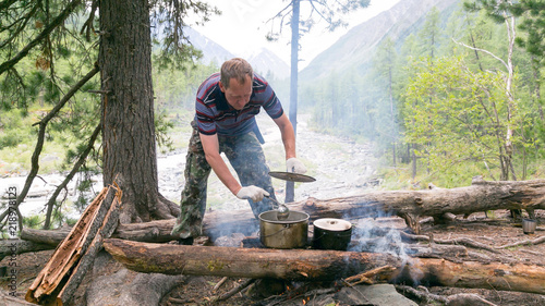 The man is cooking food at the stake in the forest. The tourist interferes with a soup sauce in a saucepan on a ksotra in a hike.