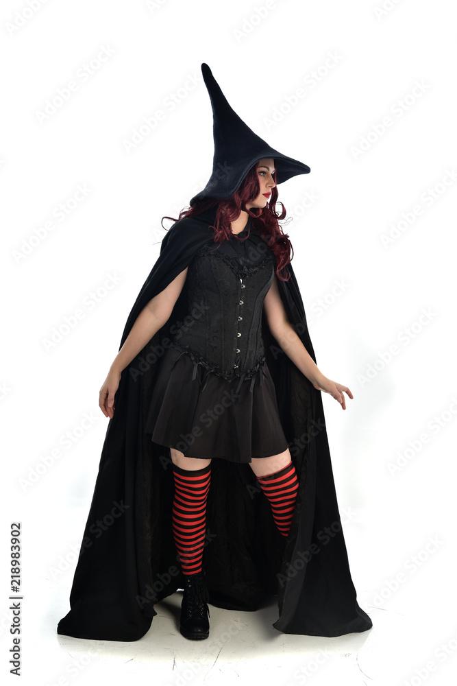 full length portrait of red haired girl wearing long black cloak, pointy hat and witch costume. standing pose, isolated on white studio background. full length portrait of red haired girl wearing long black cloak, pointy hat and witch costume. standing pose, isolated on white studio background.