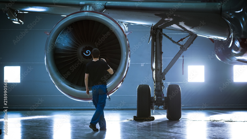 Fototapeta premium In a Hangar Aircraft Maintenance Engineer/ Technician/ Mechanic Inspects with a Flashlight Airplane's Jet Engine.