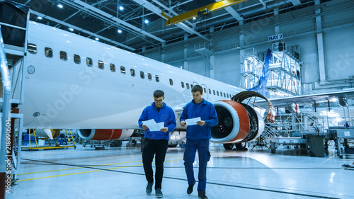 Two aircraft maintenance mechanics have a conversation while checking documents in a plane hangar with an airplane in the background. photo