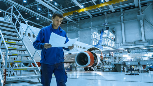 Aircraft maintenance mechanic in blue uniform is going down the stairs while reading papers in a hangar.