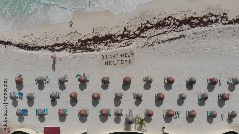 Aerial shot rising up from bienvenidos welcome sign written in the sand. On a tropical white sand beach with beach umbrellas, a message welcomes tourists and travelers in Cancun, Mexico.