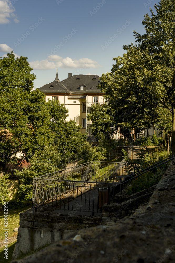 Obraz premium Stadtvilla bei strahlendem Sonnenschein an einem Park mit vielen Bäumen und alter Stadtmauer, City villa in bright sunshine at a park with many trees and old city walls