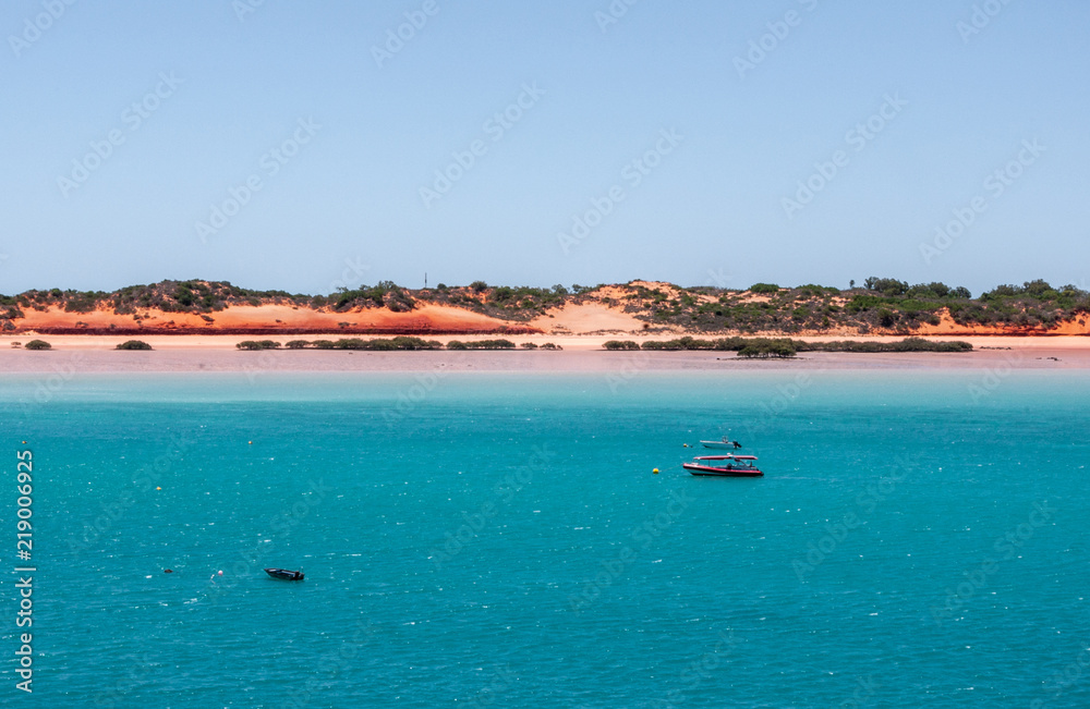 Broome, WA, Australia - November 29, 2009: Horizontal line made by red dunes with some green vegetation on top separates blue sky from azure water. Small red boat.