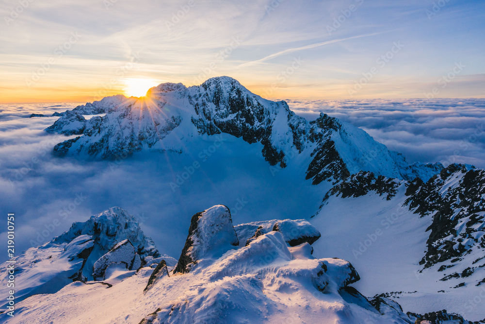 Stunning sunset or sunrise in winter alpine like snow landscape. Inversion, sun star peaking behind high rocky and icy summit. Purple, pink, blue and orange colors. Ladovy stit in winter High Tatras.