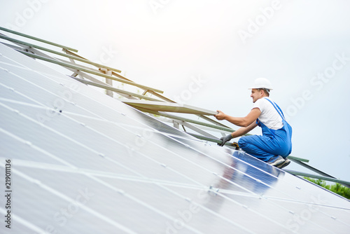 Installing of solar photo voltaic panel system. Professional worker in hard-hat and blue overall lifting the solar module on metal platform. Alternative energy and professional construction concept.