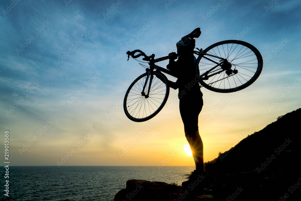 Fototapeta premium Silhouette of bicyclist carrying his bicycle on shoulder while walk against on colorful sunset orange sky background. Active outdoors lifestyle for healthy concept.
