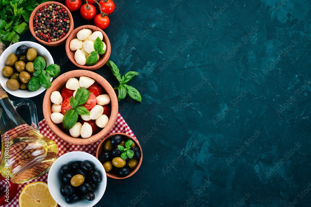 Cooking a caprese salad. Mozzarella cheese, cherry tomatoes, olives, basil leaves, oil, pepper. On a black stone table. Free space for text.