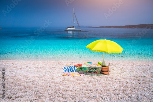 Idyllic white beach with green yellow umbrella on lazy summer day. Sailing boat at anchor in calm crystal clear blue sea water photo