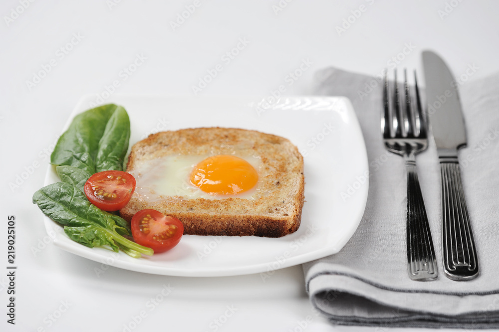 Toast with fried egg on a white plate. On a plate, spinach leaves and cherry tomatoes. Next to the dish there is a napkin and cutlery. White background. Close-up. 