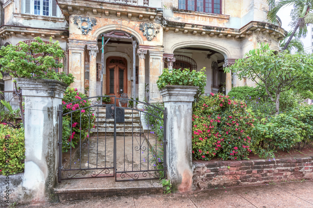 fence with bushes and a metal gate to a dilapidated old mansion with columns