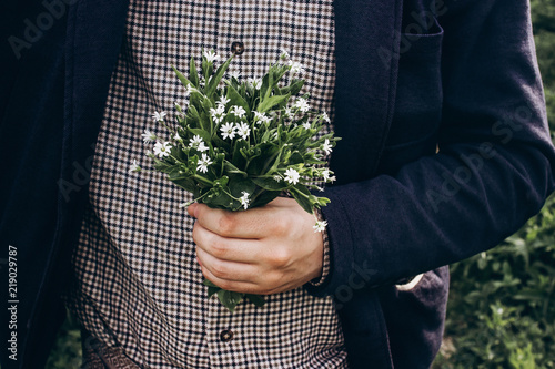 stylish hipster man holding spring bouquet of wildflowers in sunny countryside. space for text. rustic groom with simple flowers in hands. beautiful moment. save environment.