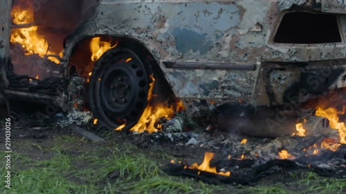 The burning car is in the field, the blown up car is on fire, the car is burning in slow motion