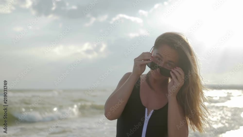 Portrait of a young brunette girl with loose hair in sunglasses and a swimsuit who walks on the beach and enjoy the sea, she looks at the horizon. On it blowing in the breeze Portrait of a young brunette girl with loose hair in sunglasses and a swimsuit who walks on the beach and enjoy the sea, she looks at the horizon. On it blowing in the breeze