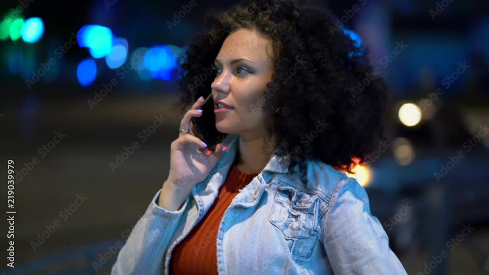 Young beautiful woman with very curly afro hair talking on her mobile phone at night illuminated street. Trendy girl have conversation. Smartphone, technology concept. 4k footage Young beautiful woman with very curly afro hair talking on her mobile phone at night illuminated street. Trendy girl have conversation. Smartphone, technology concept. 4k footage