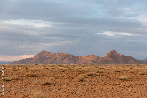 Beautiful landscape photograph of mountains in namib desert with grass foreground and cloudscape at sunrise, the sun just touching the peaks, Namibia