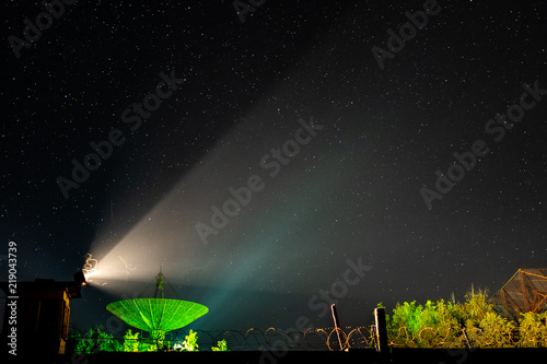 Beam of an old searchlight and line of barbed wire against the background of the big antenna of the radio telescope illuminated by green light under the night star sky.