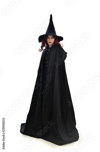 full length portrait of red haired girl wearing long black cloak, pointy hat and witch costume. standing pose, isolated on white studio background.