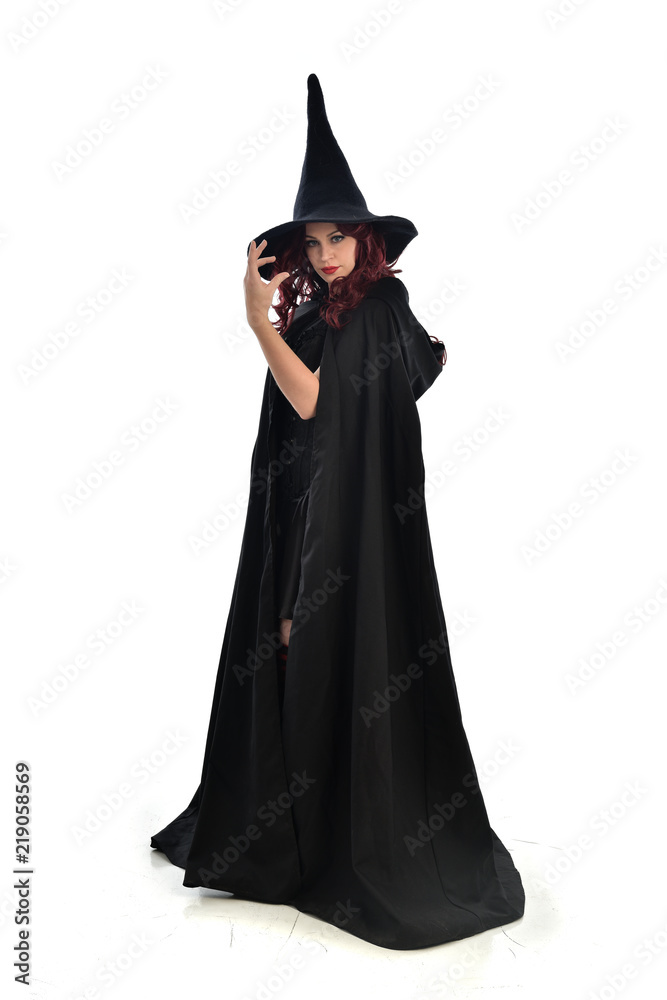 full length portrait of red haired girl wearing long black cloak, pointy hat and witch costume. standing pose, isolated on white studio background. full length portrait of red haired girl wearing long black cloak, pointy hat and witch costume. standing pose, isolated on white studio background.