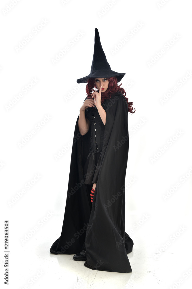 full length portrait of red haired girl wearing long black cloak, pointy hat and witch costume. standing pose, isolated on white studio background. full length portrait of red haired girl wearing long black cloak, pointy hat and witch costume. standing pose, isolated on white studio background.