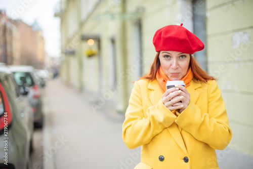 the red-haired young woman in a yellow coat and red beret is drinking coffee on the street to keep warm. A girl in bright outer clothes drinks hot tea with delight