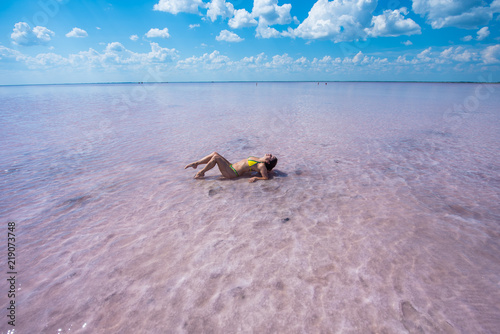 Fototapeta Naklejka Na Ścianę i Meble -  A girl in a yellow bikini is lying on her back in a pink salt lake. the bottom of the lake is covered with salt. the lake is pink because of the red algae. The girl lies on her back, leaning on her el